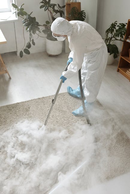 A professional cleaner dressed in a full white protective suit, gloves, and face mask is performing deep cleaning on a light-colored, plush carpet in a well-lit, modern living room. The cleaner is usi