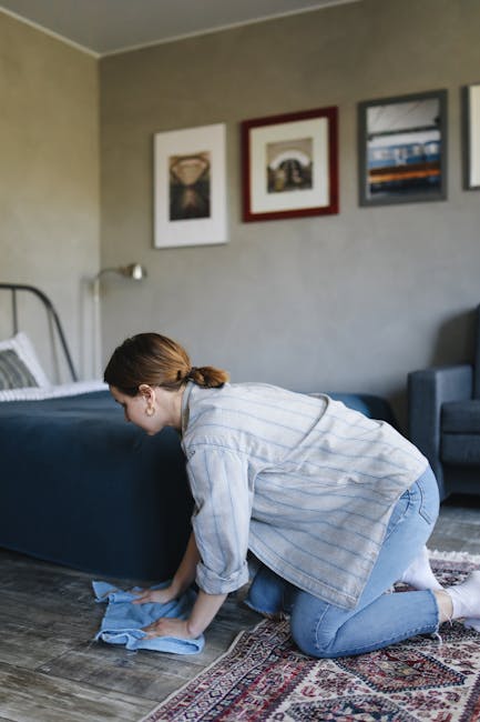 A professional cleaner dressed in a full white protective suit, gloves, and face mask is performing deep cleaning on a light-colored, plush carpet in a well-lit, modern living room. The cleaner is usi