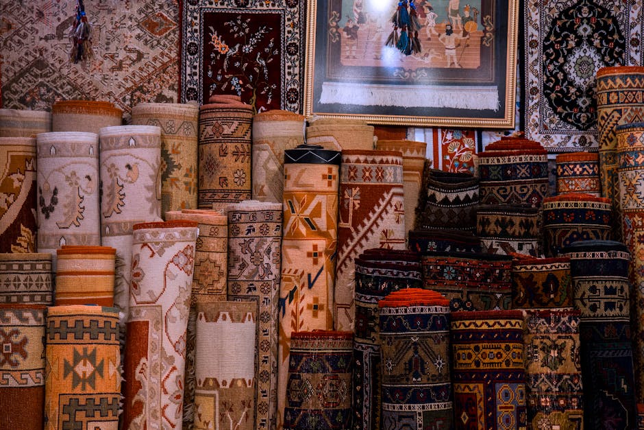 An indoor textile shop displaying a variety of traditional and decorative rugs, with some rolled and stacked on the floor and others hanging on the walls. The rugs feature intricate patterns and vibra